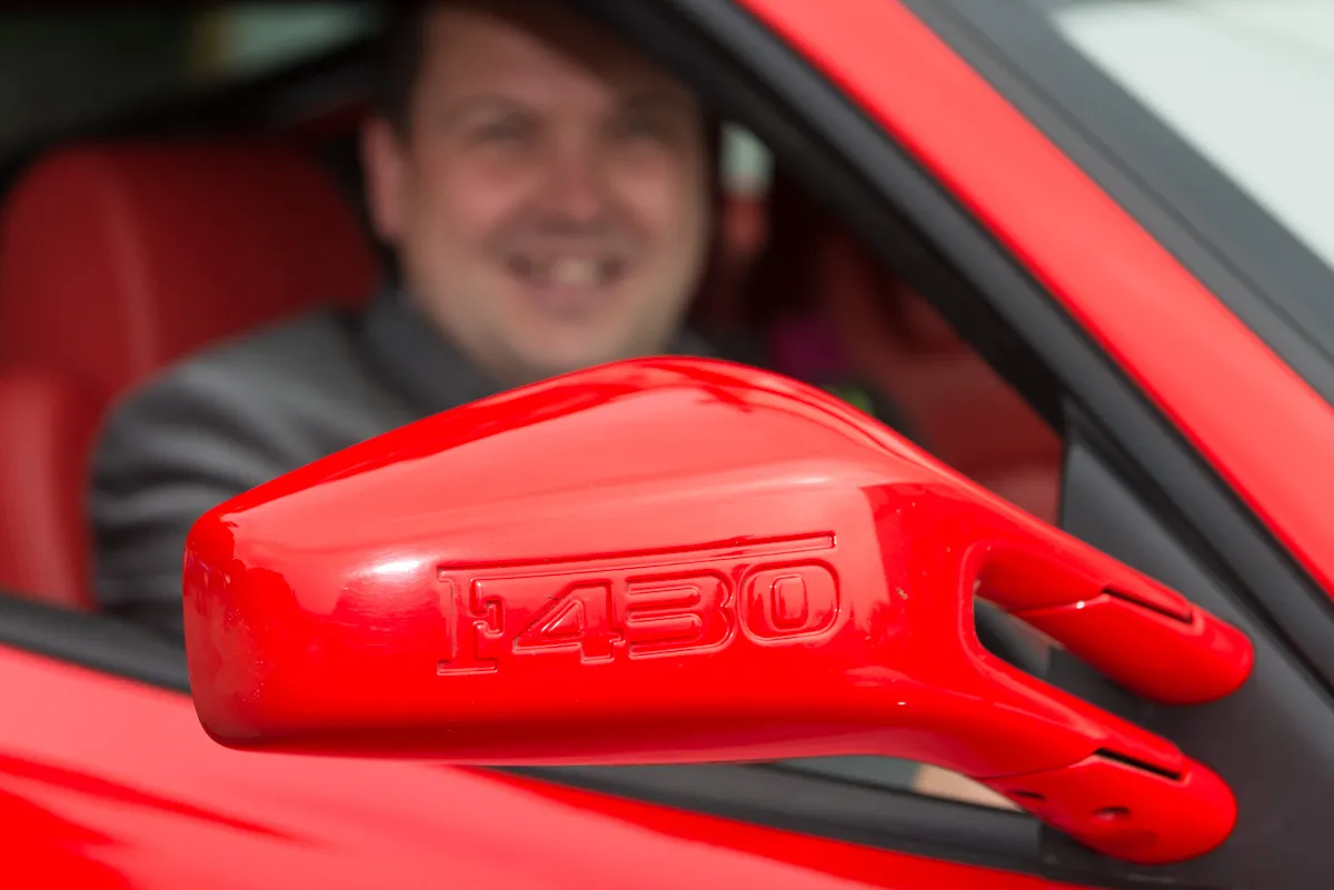Close-up of a red car's side mirror with 'F430' embossed on it and a blurred smiling man sitting inside.