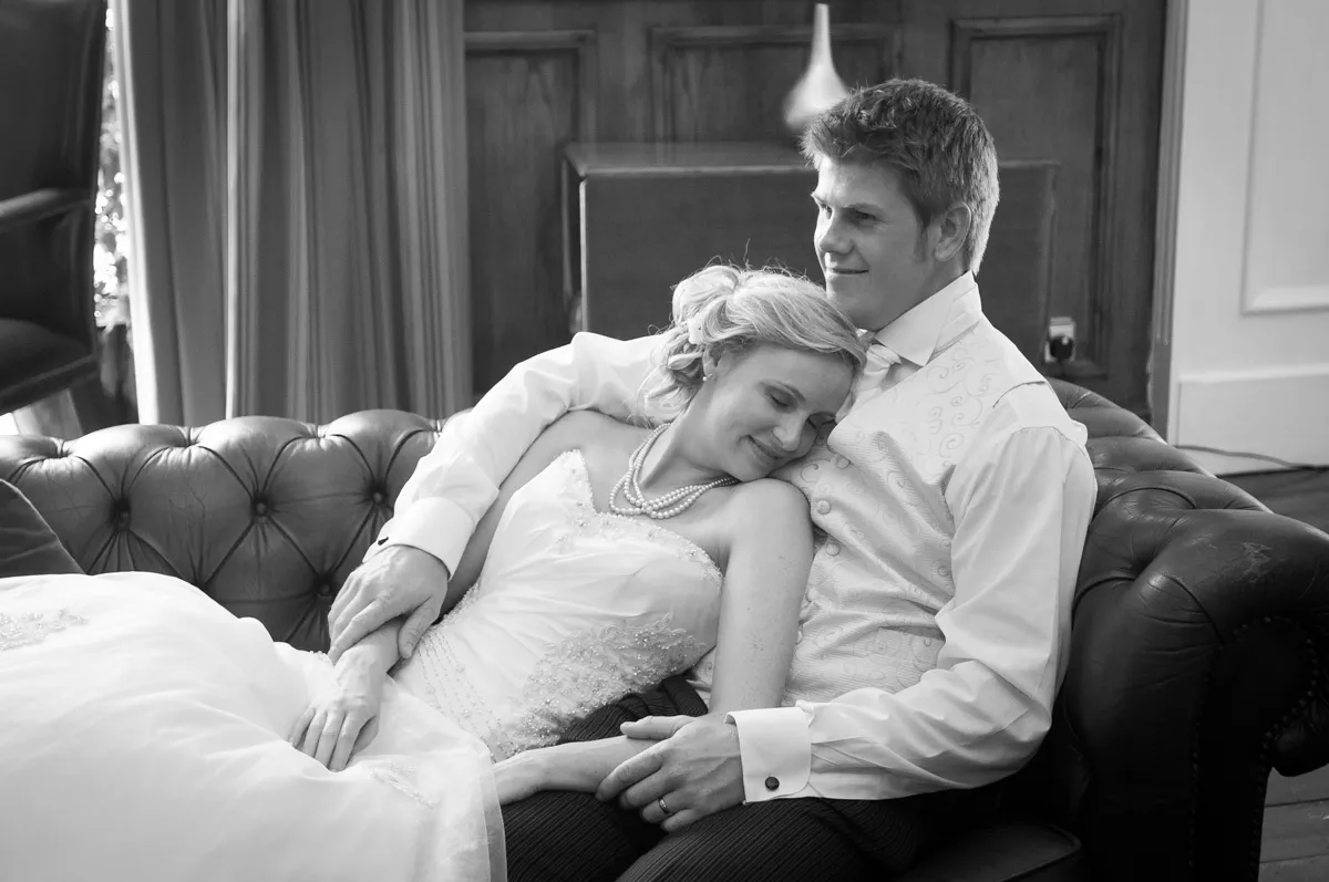 Black-and-white photo of a bride and groom sitting and cuddling on a leather sofa, the bride resting her head on the groom's chest.