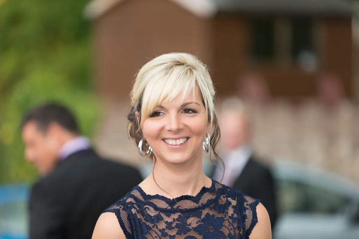 Smiling blonde woman with silver hoop earrings wearing a dark lace dress in an outdoor setting.