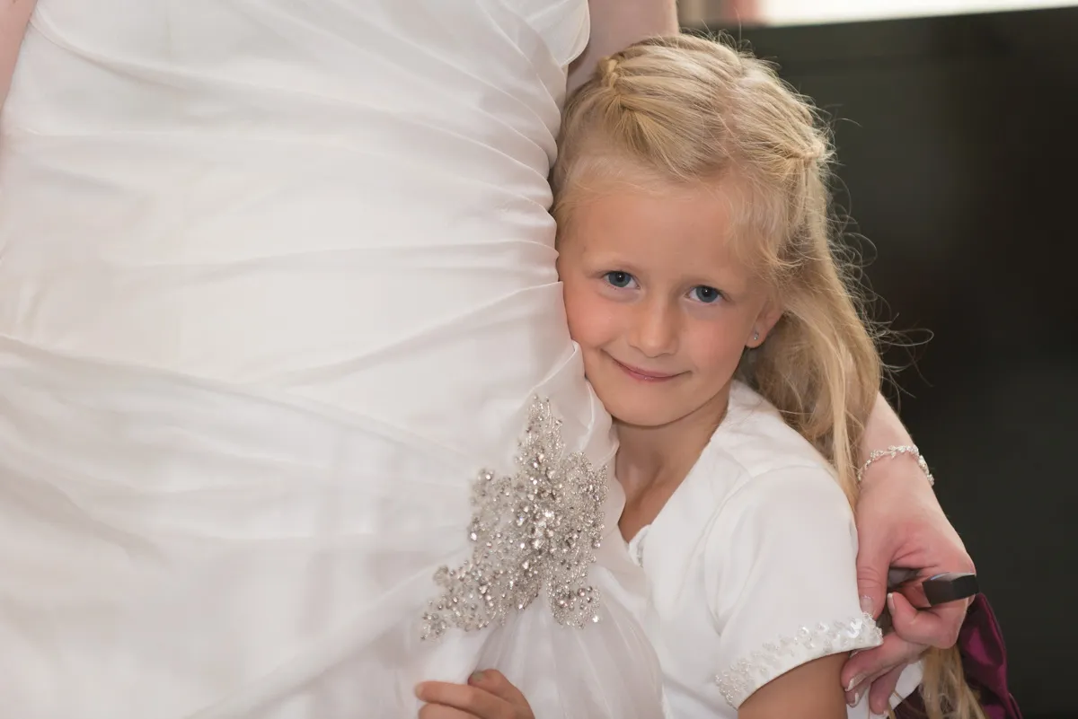 Young girl with blonde hair hugging the waist of a bride in a white wedding dress.
