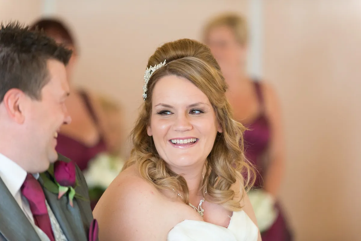 Smiling bride with styled blonde hair and hair accessory, looking at groom in gray suit and burgundy tie.