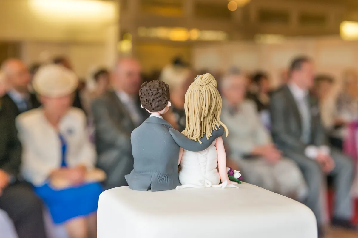 Wedding cake topper of a bride and groom sitting side by side with the groom's arm around the bride, with blurred guests in the background.