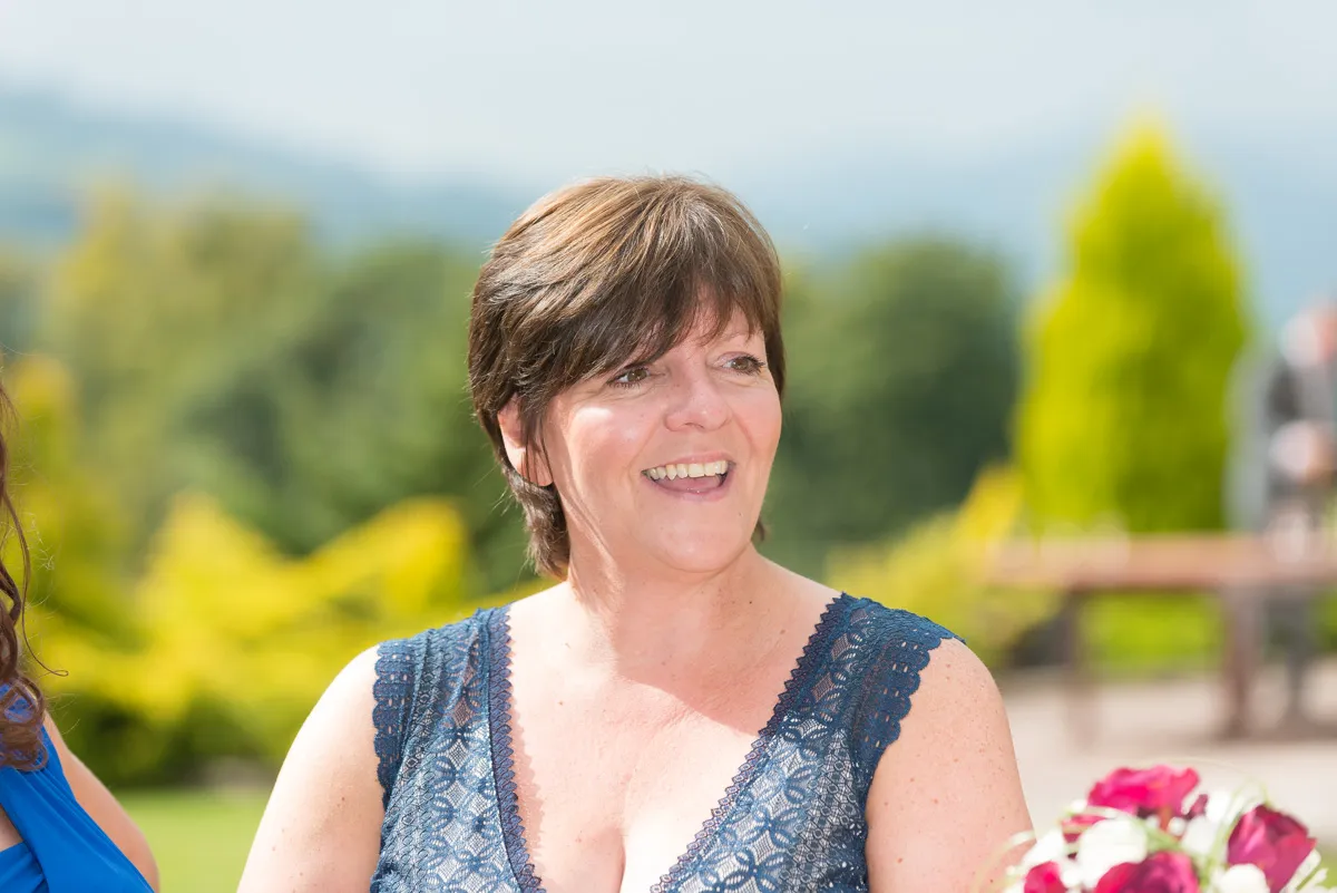 Smiling woman with short brown hair wearing a blue lace dress, outdoors in a garden setting.