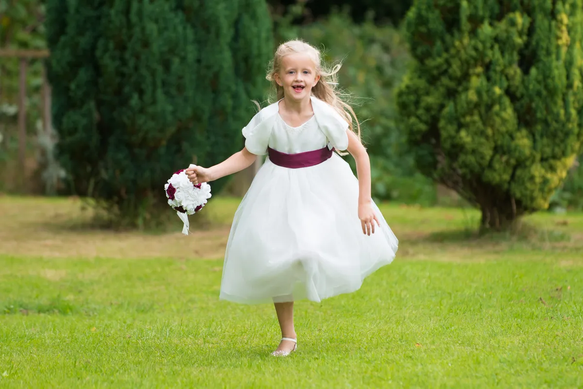 Young girl in a white dress with a purple sash running on grass holding a bouquet of white and purple flowers.