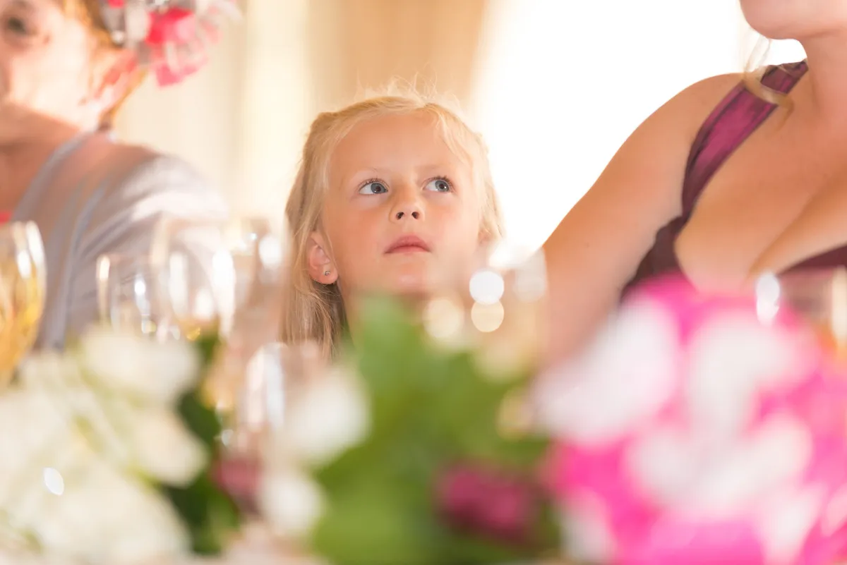 Young girl with blonde hair looking upward, seated at a table with blurred flowers and glasses in the foreground.