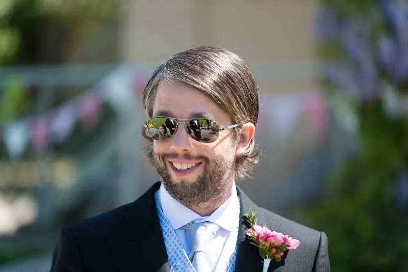 Smiling man in a black suit, sunglasses, white shirt, and tie with a pink boutonniere.