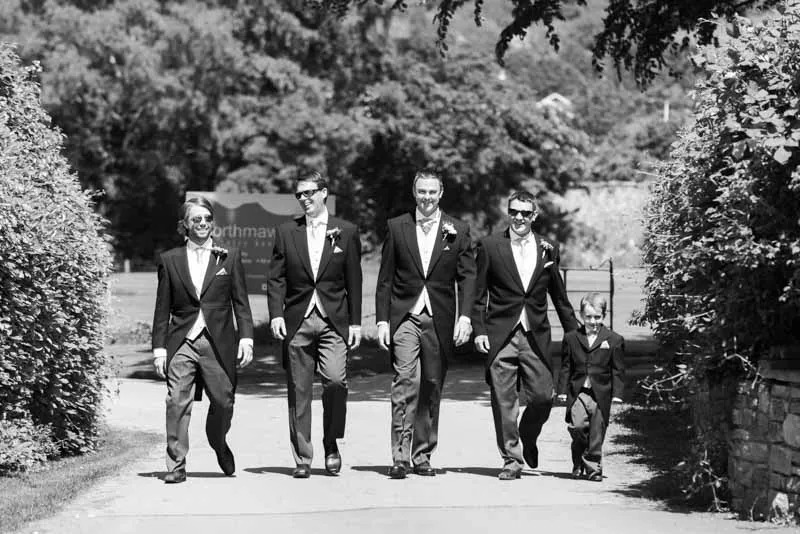 Five males dressed in tuxedos walking on a sunlit path outdoors with greenery on both sides.