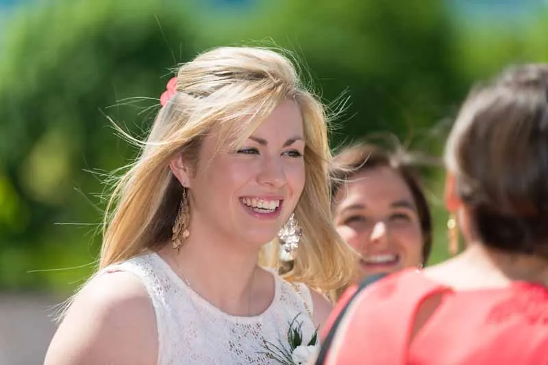 Smiling blonde woman in a white lace dress outdoors, with two other people partially visible and blurred in the background.