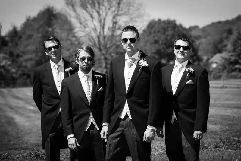 Four men in formal suits and sunglasses standing outdoors on grass with trees and hills in the background.