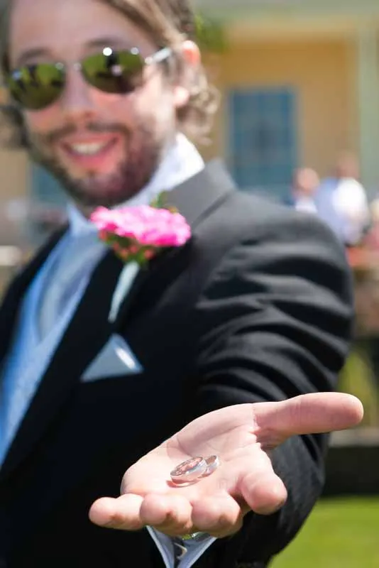 Man in a suit with a pink boutonniere holding two wedding rings in his open palm outdoors.