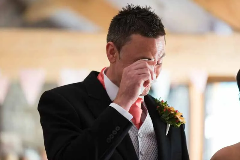 Man in formal suit wiping tears from his eyes during a ceremony, wearing a boutonniere and coral tie.