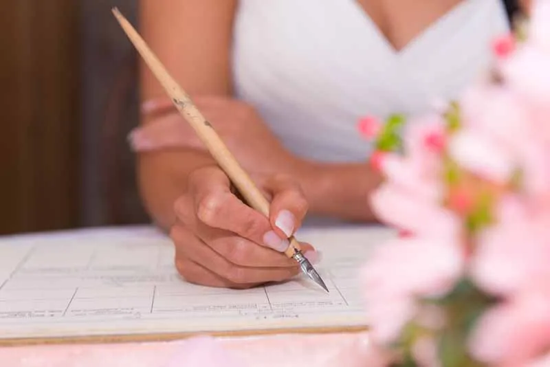 Person holding a calligraphy pen writing on a document, with blurred pink flowers in foreground.