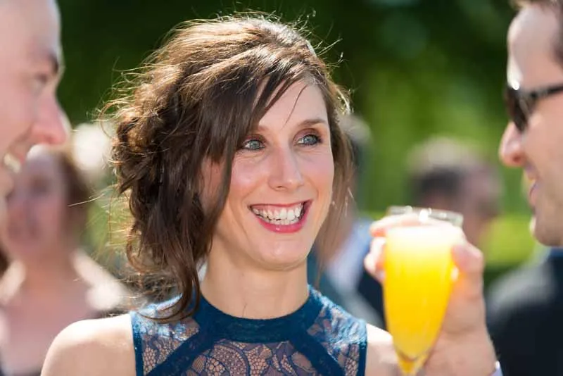 Smiling woman in a blue lace dress holding a yellow drink, engaging with two people in an outdoor setting.