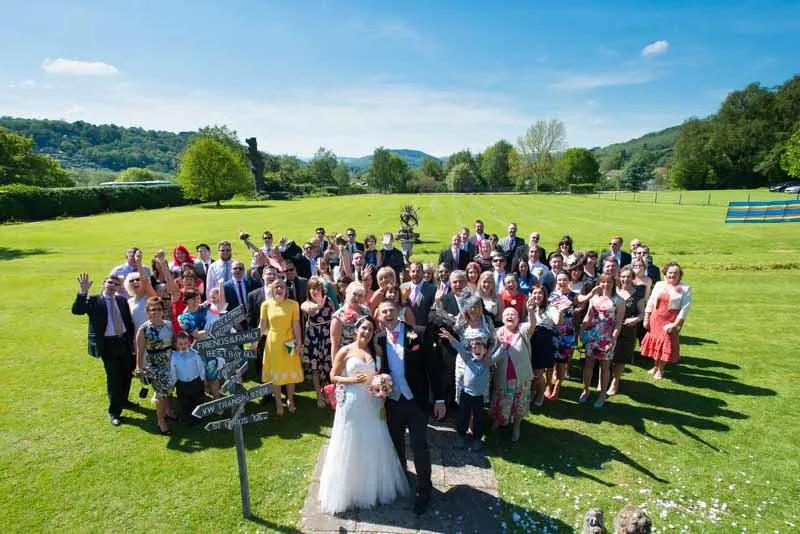 Group of wedding guests posing outdoors on a sunny day with the bride and groom at the front on a green lawn.
