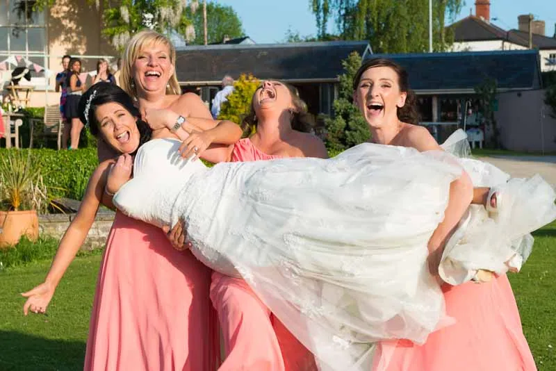 Four women laughing outdoors, three in pink dresses lifting a woman in a white wedding gown.