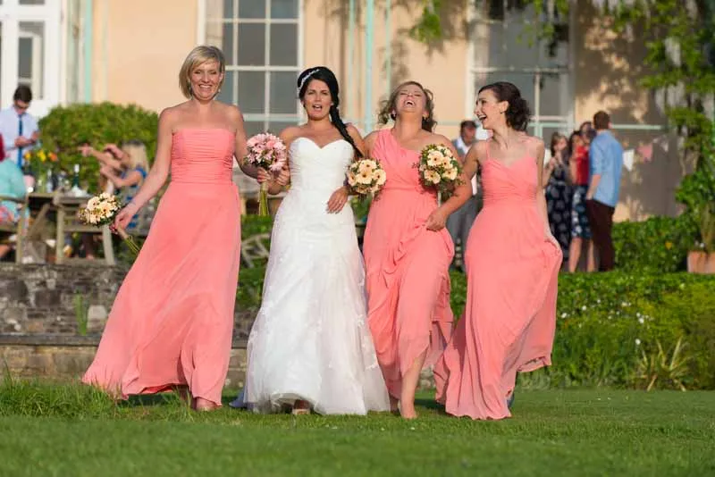 Bride in white dress walking arm-in-arm with three bridesmaids in coral dresses on a grassy lawn.