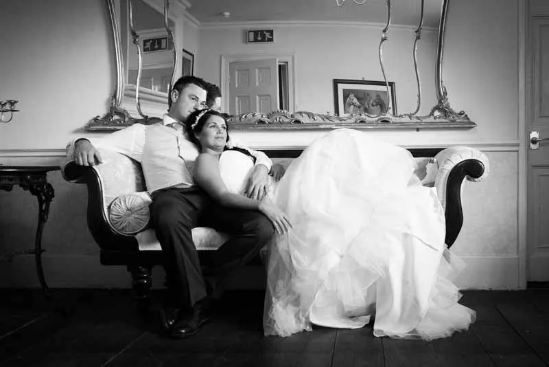 Bride in wedding dress reclining on a sofa with groom in vest and tie sitting beside her in a classic room with ornate mirror and framed artwork.