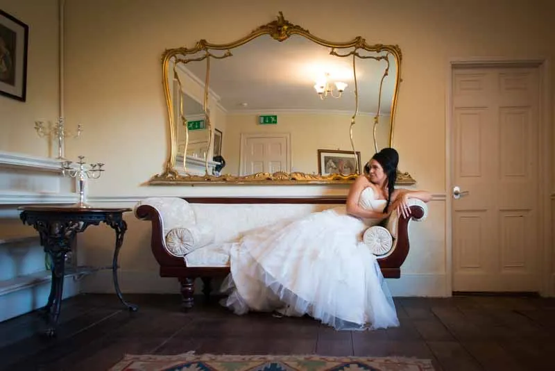 Bride in a white wedding dress lounging on a vintage wooden sofa with ornate gold mirror behind her.
