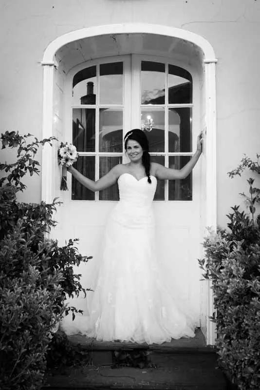 Bride in a strapless wedding gown holding a bouquet standing in a doorway framed by bushes.