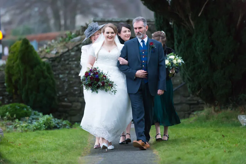 Bride in white wedding dress holding a bouquet, walking arm-in-arm with a man in a suit, followed by bridesmaids on a garden path.