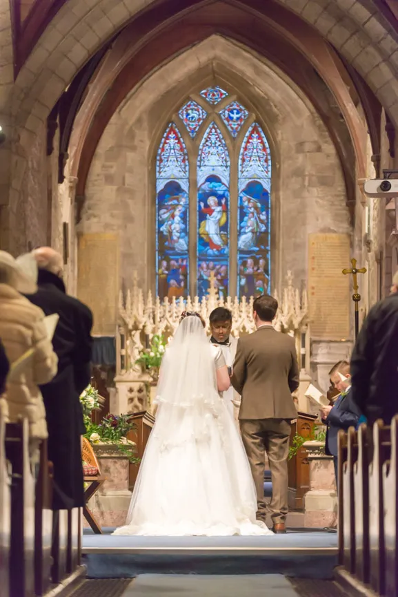 Bride and groom standing at the altar in a church during a wedding ceremony with stained glass window in the background.