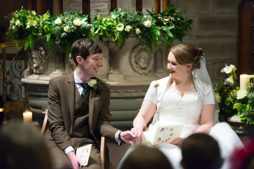 Bride and groom holding hands and smiling at each other during a wedding ceremony in a decorated church setting.