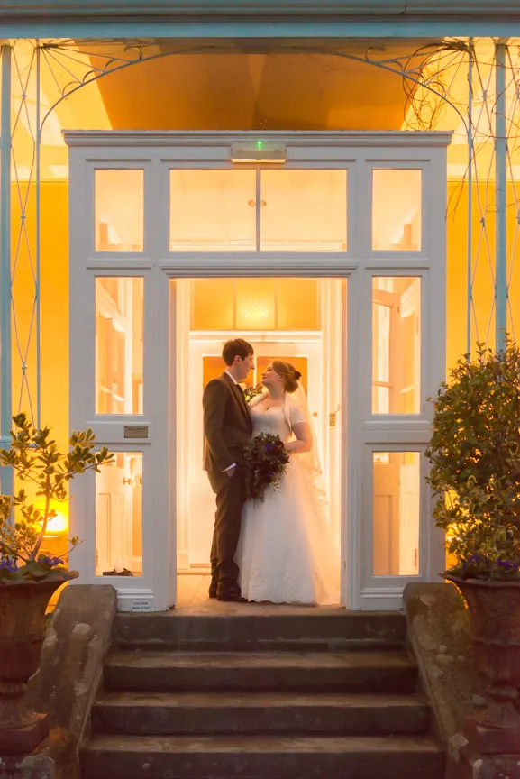 Bride and groom standing face to face at the lit entrance of a building in the evening.