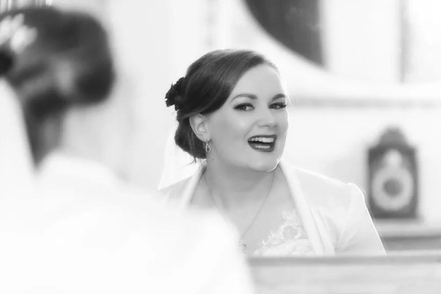 Smiling bride with styled hair and veil seen in a mirror, wearing a lace wedding dress and earrings.