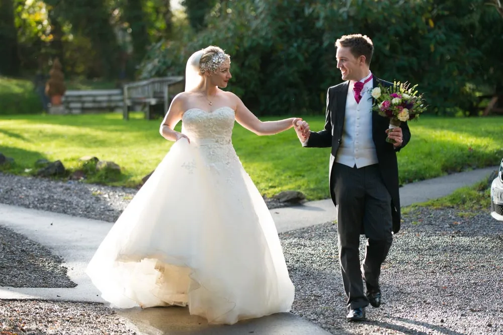 Bride and groom holding hands and smiling, walking outdoors on a gravel path with green grass and trees in the background.