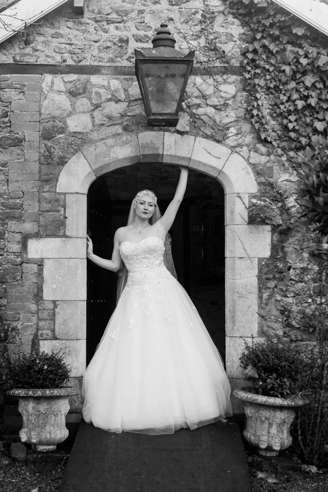 Bride in a strapless lace wedding gown and veil standing under a stone archway with one arm raised touching a hanging lantern.
