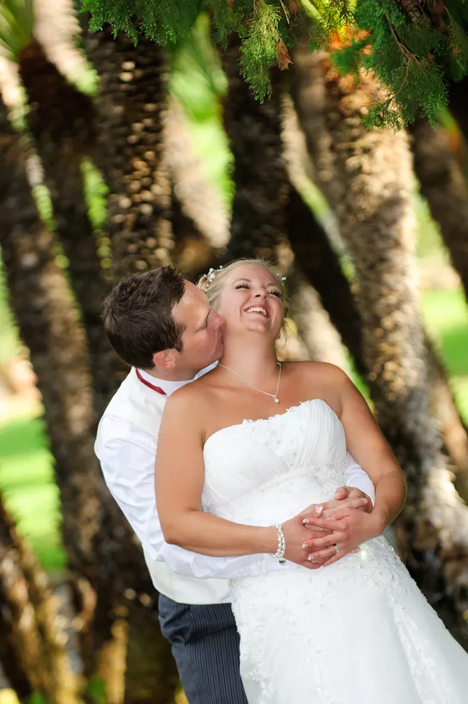 Bride in white dress embraced and kissed on the cheek by groom in white shirt against backdrop of palm trees.
