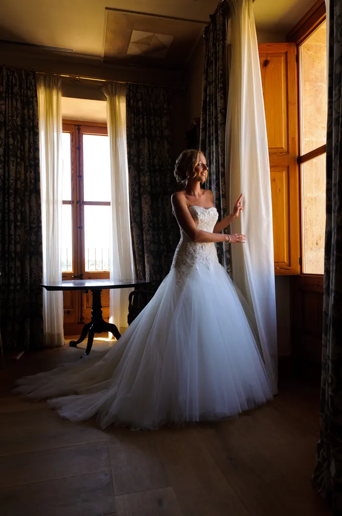 Bride in a white strapless wedding dress standing by a wooden window with sheer and patterned curtains.