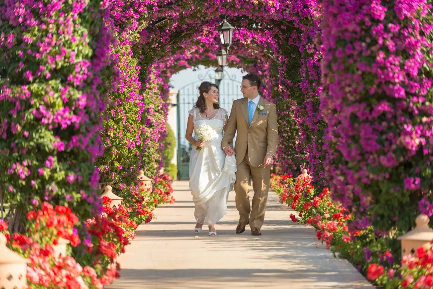Bride and groom walking hand in hand under an archway covered with vibrant pink and red flowers.