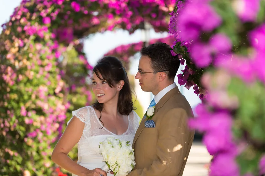 Bride in white holding bouquet and groom in brown suit with blue tie standing under archway of pink flowers.