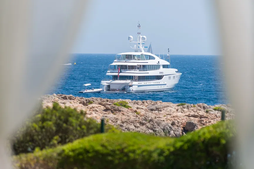 White luxury yacht anchored near rocky shore with blue sea under clear sky.