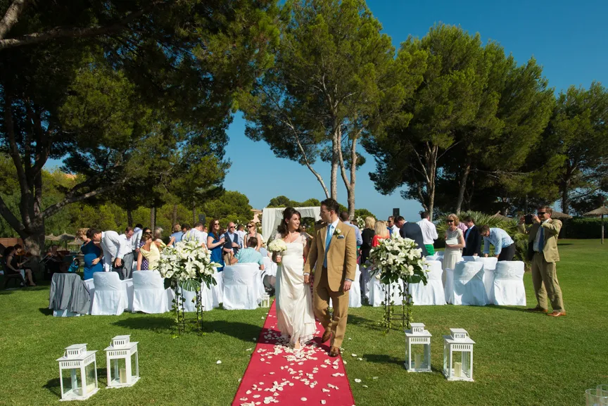 Bride and groom walking down a red aisle outdoors with guests seated on white-covered chairs under trees on a sunny day.