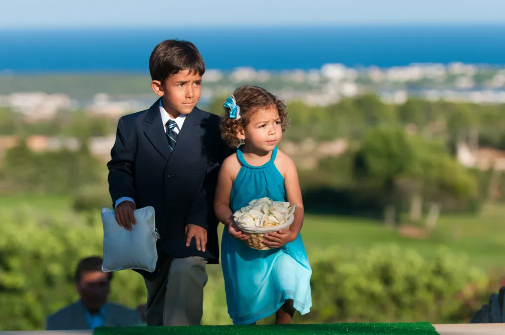 A young boy in a suit holding a white pillow and a young girl in a blue dress holding a basket of white rose petals outdoors with a blurred background of green landscape and blue sky.