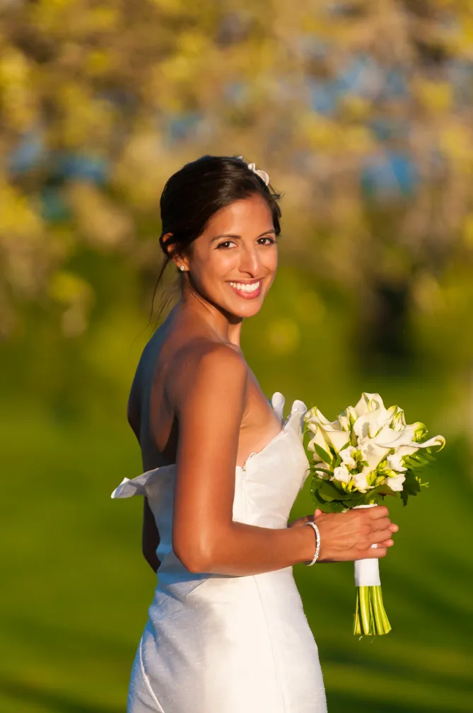 Smiling bride in a strapless white wedding dress holding a bouquet of white flowers against a blurred outdoor background.