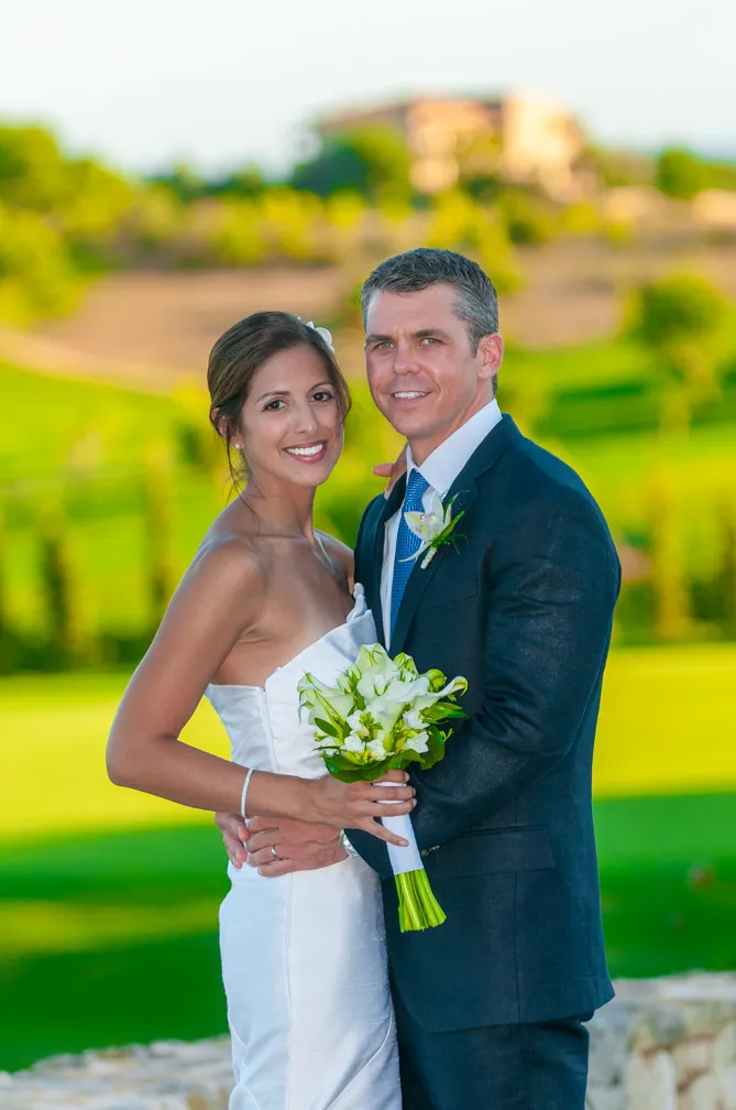 Bride in a white strapless dress holding a bouquet and groom in a dark suit posing outdoors on their wedding day.