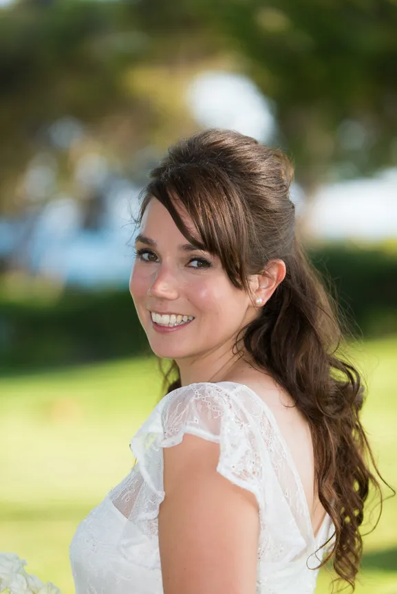 Smiling bride with long brown hair in a white lace wedding dress outdoors.