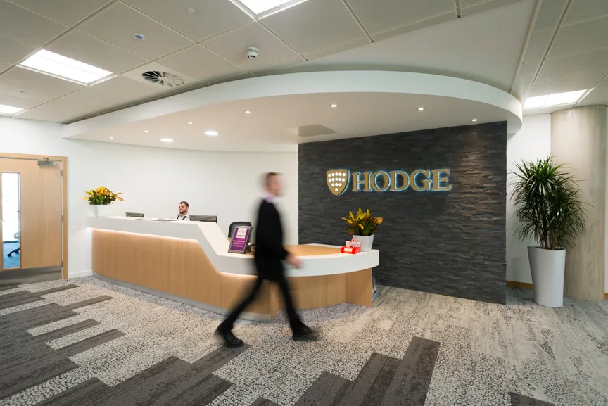 Reception area with a wooden desk, a person walking blurred past, receptionist seated, and a dark stone wall with illuminated Hodge logo.