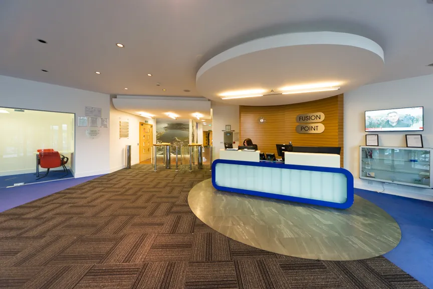 Modern office reception area with a circular overhead light fixture, a white desk with blue trim, and a wooden wall sign reading 'Fusion Point'.