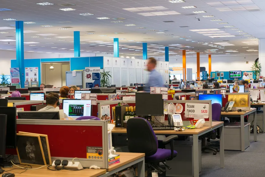 Large open office space with rows of desks, computer monitors, and people working in cubicles under bright fluorescent lighting.