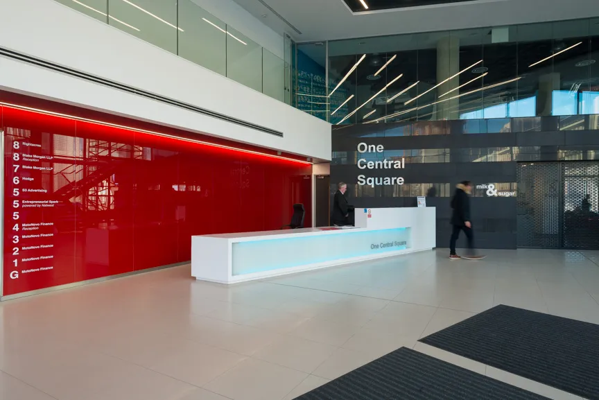 Modern office lobby with a white reception desk, red directory wall, and black tiled wall displaying 'One Central Square'.