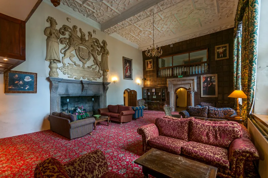 Ornate historical sitting room with patterned sofas, a large stone fireplace, decorative ceiling, wood paneling, and classic paintings.