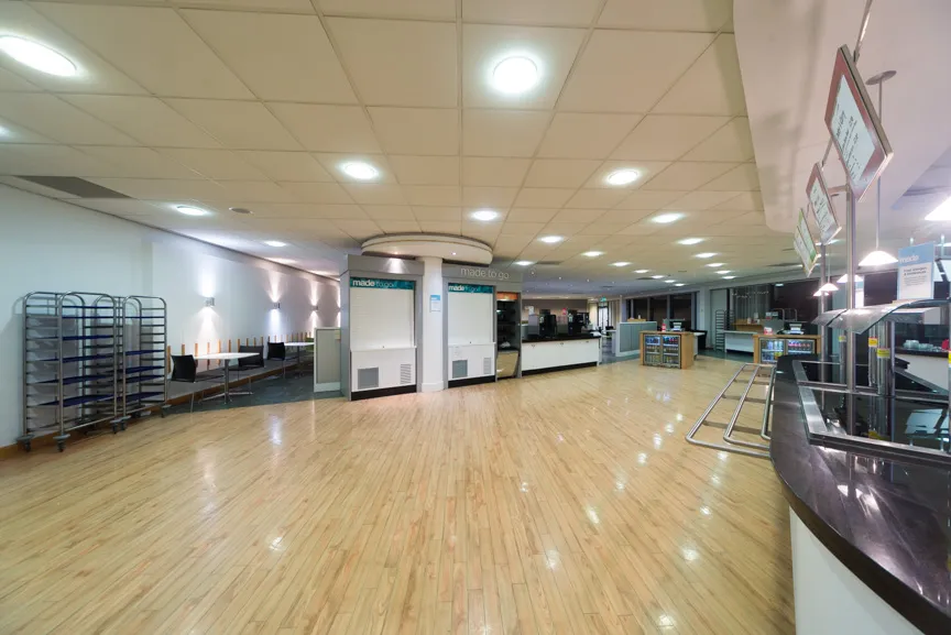 Empty cafeteria with wooden floor, ceiling lights, closed food service counters, and stacked trays on the left side.