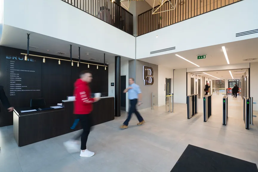 Modern office lobby with a reception desk, glass entrance gates, and people walking inside.