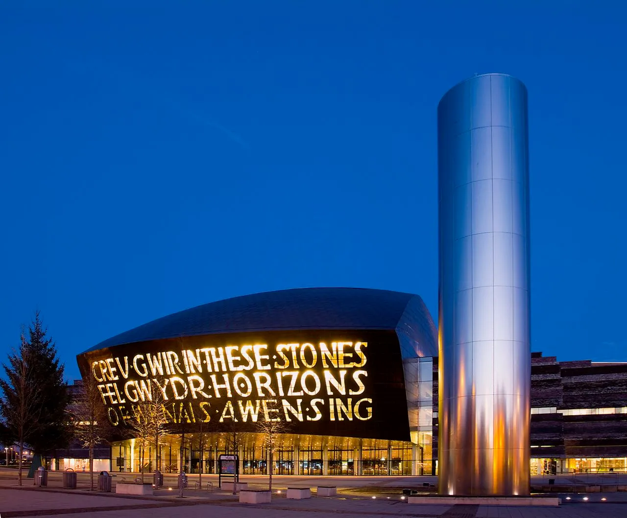 Cardiff's Wales Millennium Centre at dusk with illuminated Welsh and English text on its curved frontage and a tall metallic cylindrical sculpture in the foreground.