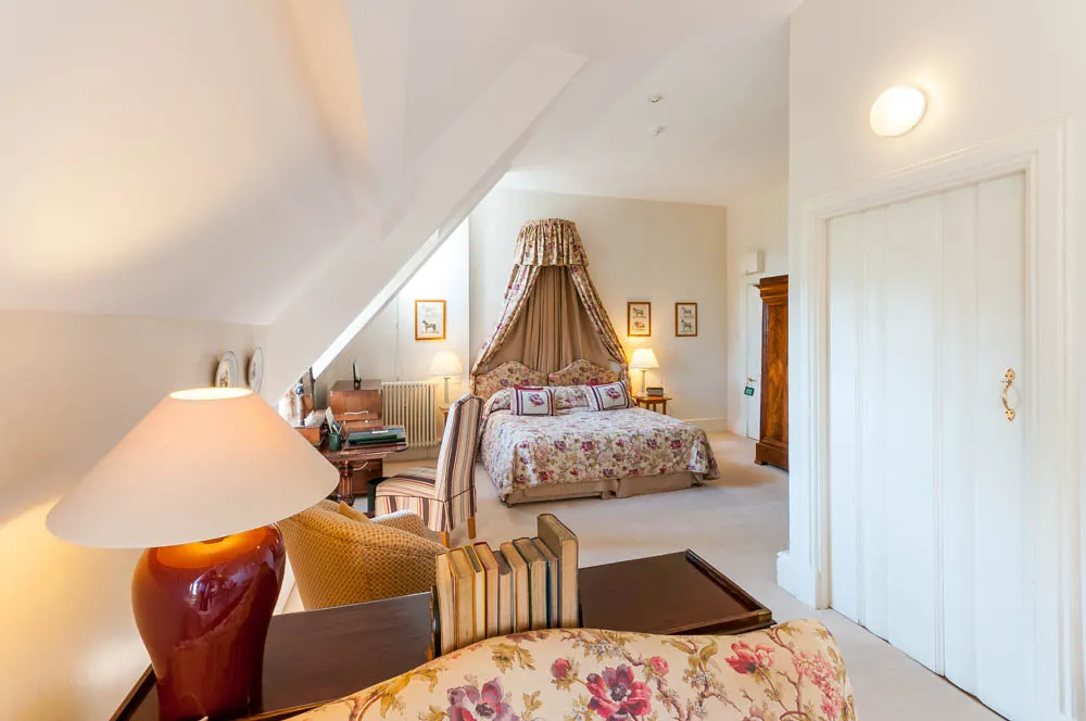 Bright attic bedroom with floral patterned bedspread under a fabric canopy, wooden furniture, a desk with books, and a red table lamp.