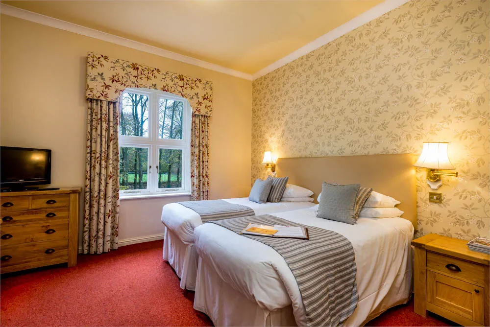 Cozy hotel room with two twin beds, floral wallpaper, a window with floral curtains, wooden dresser, and bedside table with lamps.